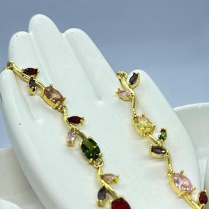 May include: A gold-tone metal bracelet featuring a vine-like design adorned with multi-colored, oval and marquise-cut gemstones. The stones include shades of red, green, yellow, pink, and purple, arranged along the flexible, branching metal links.