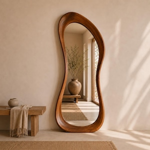 May include: A full-length floor mirror featuring a thick, organic, asymmetrical wooden frame with a smooth, polished finish. The mirror reflects a bright interior space containing a wooden bench, a ceramic vase, and a woven rug on the floor.