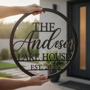 Könnte beinhalten: Ein kreisförmiges schwarzes Metallschild mit der Aufschrift THE Anderson LAKE HOUSE EST. 2023. Das Schild hat ein klares Design mit einer fetten serifenlosen Schrift für die Wörter THE, LAKE HOUSE und EST. 2023 sowie einer Schreibschrift für Anderson.