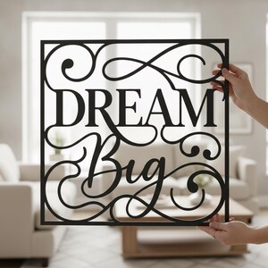 Könnte beinhalten: Ein quadratisches Wandbild aus schwarzem Metall mit dem Schriftzug "DREAM Big" in einer Kombination aus Serifenschrift und Schreibschrift, umgeben von dekorativen, geschwungenen Filigranmustern, die mit dem Außenrahmen verbunden sind.