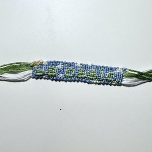 May include: A handmade woven friendship bracelet featuring a blue background with white star patterns and the word "so basic" spelled out in light green thread. The bracelet ends in loose, frayed green and white embroidery floss tassels.