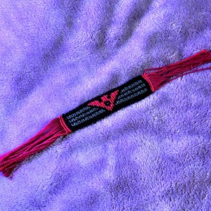 May include: A handmade woven friendship bracelet featuring a black background with a central pink geometric bird-like emblem. The bracelet is crafted from thread and includes long, loose pink fringe ends extending from both sides of the woven section.