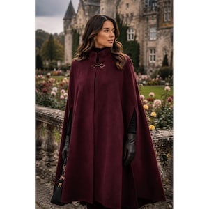 May include: A deep burgundy wool-blend cape featuring a high collar and a metallic toggle clasp closure at the neck. The garment has arm slits on the sides and a flowing, floor-length silhouette, worn over a black long-sleeve top and black gloves.