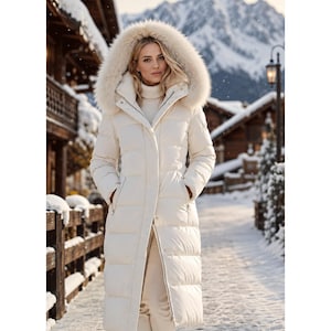 May include: A long, cream-colored puffer coat featuring a high collar, a full-length front zipper with a snap-button placket, and a large, plush faux fur-trimmed hood. The coat has a quilted, horizontal-baffled design and side pockets.