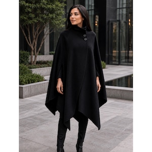 May include: A black wool-blend cape featuring a high collar and two large circular buttons at the neckline. The garment has an asymmetrical hemline, a structured silhouette, and wide arm openings, providing a draped, flowing appearance.