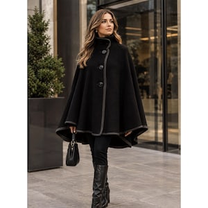 May include: A black wool-blend cape coat featuring a high collar, three large round buttons down the front, and a contrasting grey trim along the edges. The garment has a flared silhouette and is paired with black trousers and black leather boots.
