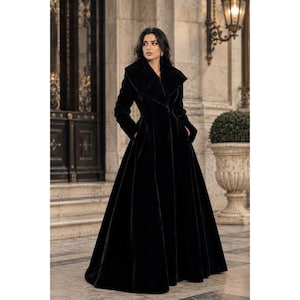 Può includere: Un cappotto lungo fino ai piedi in velluto nero caratterizzato da un ampio colletto oversize, maniche lunghe e un corpetto aderente che si apre in una gonna ampia e voluminosa. Il cappotto è realizzato in tessuto nero tinta unita e morbido.