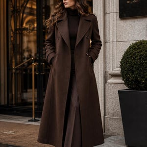 K&ouml;nnte beinhalten: Ein langer, dunkelbrauner zweireihiger Wollmantel mit breitem Revers und Seitentaschen, getragen &uuml;ber einem passenden dunkelbraunen gerippten Rollkragenpullover und einer weiten Hose. Der Mantel hat strukturierte Schultern und eine lange Form.