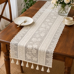 Puede incluir: Un camino de mesa rectangular de encaje en color blanco roto, con un panel central de motivos florales flanqueado por bordes decorativos con diseños circulares y de hojas. Los extremos están rematados con pequeñas borlas.