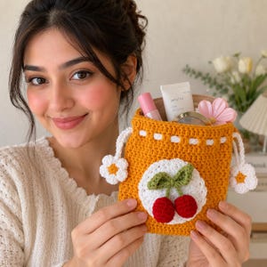 May include: A mustard yellow crochet drawstring pouch featuring a white circular patch with two red cherries and green leaves. The pouch has white crocheted flower accents on the sides and a white drawstring cord, containing various cosmetic items.