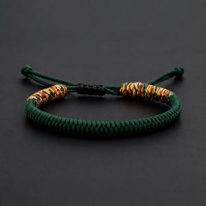 May include: A dark green braided cord bracelet featuring two decorative segments of interwoven gold, red, and dark green threads. The bracelet includes a black sliding knot closure and two thin green cord ends with knotted tips for adjustable sizing.