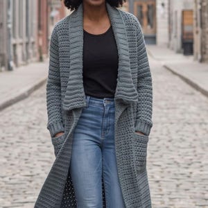 May include: A long, dark grey, open-front crochet cardigan featuring a wide shawl collar, long sleeves, and two large patch pockets. The garment is worn over a black scoop-neck top, light blue distressed denim jeans, and black ankle boots.