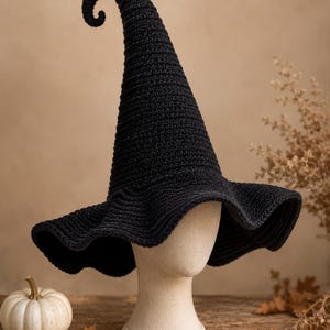 May include: A black crocheted witch hat with a tall, pointed crown that features a distinct curled tip and a wide, wavy brim. The hat is displayed on a neutral-colored mannequin head, showcasing the textured yarn construction and the structured shape.