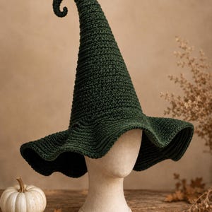 Puede incluir: Un sombrero de ganchillo en color verde bosque oscuro con una parte superior alta, puntiaguda y curvada. El sombrero presenta un ala ancha y ondulada con un patrón de punto acanalado texturizado, expuesto sobre un maniquí de color neutro.