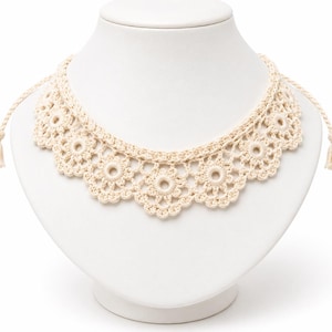 May include: A cream-colored crochet necklace featuring a repeating floral pattern with circular centers and scalloped edges. The piece includes thin braided ties on each side that terminate in small, matching decorative tassels for closure.