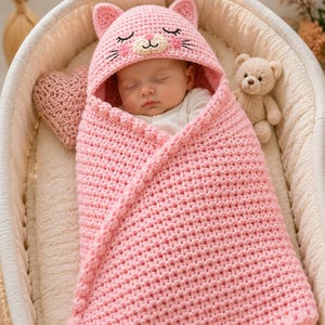 Può includere: Una coperta avvolgente per neonati lavorata all'uncinetto in rosa chiaro, dotata di cappuccio integrato con orecchie da gatto, occhi chiusi ricamati, baffi e un piccolo naso. La coperta presenta un motivo a maglia testurizzato e un bordo.