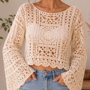 Peut inclure: Un haut à manches longues en crochet de couleur crème présentant un motif ajouré complexe avec un motif central de style mandala, une large encolure bateau et des manches évasées, porté avec un jean en denim bleu clair.