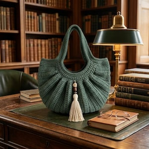 May include: A dark green crochet handbag with a pleated, fan-like texture and a rounded silhouette. The bag features a central decorative accent consisting of a dark wooden bead, a light wooden bead, and a long, cream-coloured fabric tassel.