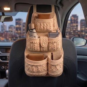 May include: A beige crocheted car seat organizer featuring multiple pockets with cable knit and bobble textures. The organizer is attached to the back of a car seat and holds a brown wallet, a folded umbrella, a power bank, a notebook, and a pen.