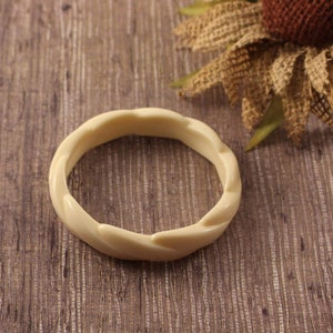 Peut inclure: Un bracelet jonc de couleur crème présentant un motif torsadé ou en forme de corde continue. L'accessoire circulaire possède une finition lisse et polie ainsi qu'une épaisseur uniforme sur toute sa circonférence, affichant une esthétique simple.