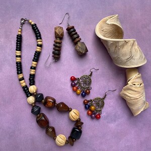 Puede incluir: Un collar y dos pares de pendientes de madera y metal. El collar presenta cuentas de madera negras, beige y marrones. Un par de pendientes tiene cuentas de madera apiladas, mientras que el otro par muestra círculos de metal con cuentas de colores.
