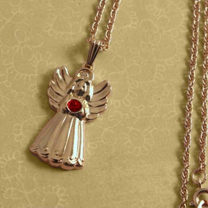 May include: A gold-colored angel pendant necklace with a red gemstone in the center. The angel has detailed wings and a flowing gown. The necklace chain is a delicate rope style.
