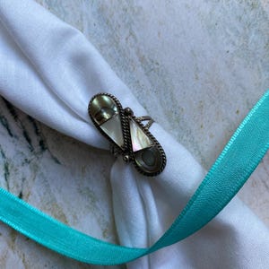 May include: A silver ring with an elongated, geometric design. The ring features mother-of-pearl inlays and a dark border. The ring is displayed on a white cloth with a turquoise ribbon.