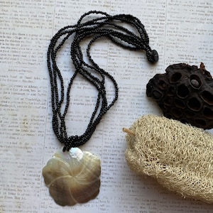 Puede incluir: Un collar de múltiples hilos con pequeñas cuentas negras y un gran colgante de concha iridiscente tallada en forma de flor. El collar incluye un pequeño cierre de botón negro en la parte posterior de los hilos de cuentas, creando un aspecto en capas.