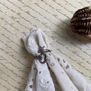 Pode incluir: Dois anéis prateados, um em forma de chave e outro de fechadura, são exibidos em tecido branco bordado com ilhós. O fundo apresenta papel vintage com escrita manuscrita e uma vagem de semente texturizada.