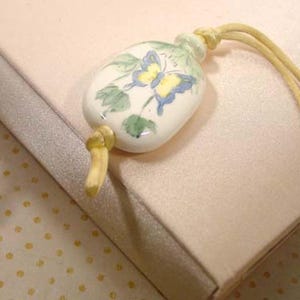 May include: A white ceramic pendant with a hand-painted butterfly design in yellow and blue, accented with green leaves. The pendant is attached to a beige cord. The pendant is approximately 3.8 cm long.