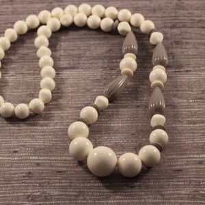 Op de afbeelding: Een ketting met een reeks gladde, ronde, gebroken witte kralen, afgewisseld met kleinere, platte, grijze tussenkralen en twee langwerpige, geribbelde, grijze kralen die asymmetrisch zijn geplaatst en eindigen in een grote centrale kraal.