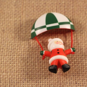 May include: A small figurine of a Santa Claus character suspended from a white and green chequered parachute. The Santa figure wears a red suit with black boots and is attached to the parachute by two thin red cords, creating a hanging ornament.