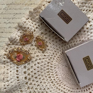 Könnte beinhalten: Eine goldfarbene Brosche und ein Paar passende ovale Ohrringe, jeweils mit einer zentralen rosa Rose auf beigem, marmoriertem Grund in einem verzierten Rahmen. Zwei graue Avon-Schachteln liegen bei dem Schmuckset, das auf einem cremefarbenen Häkelstoff liegt.