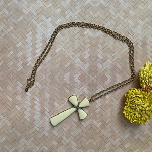 May include: A gold-tone metal chain necklace featuring a large, smooth, cream-colored cross pendant. The cross has a central decorative wrap of twisted gold-tone wire at the intersection of its arms. The necklace includes a circular spring ring clasp.