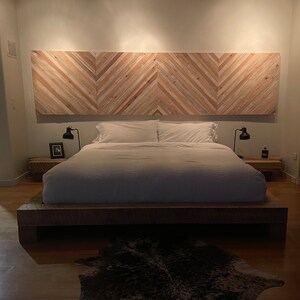 May include: A white bed with a wooden headboard made of light wood planks arranged in a chevron pattern. The bed has a white duvet and two white pillows. There are two bedside tables with lamps on either side of the bed.