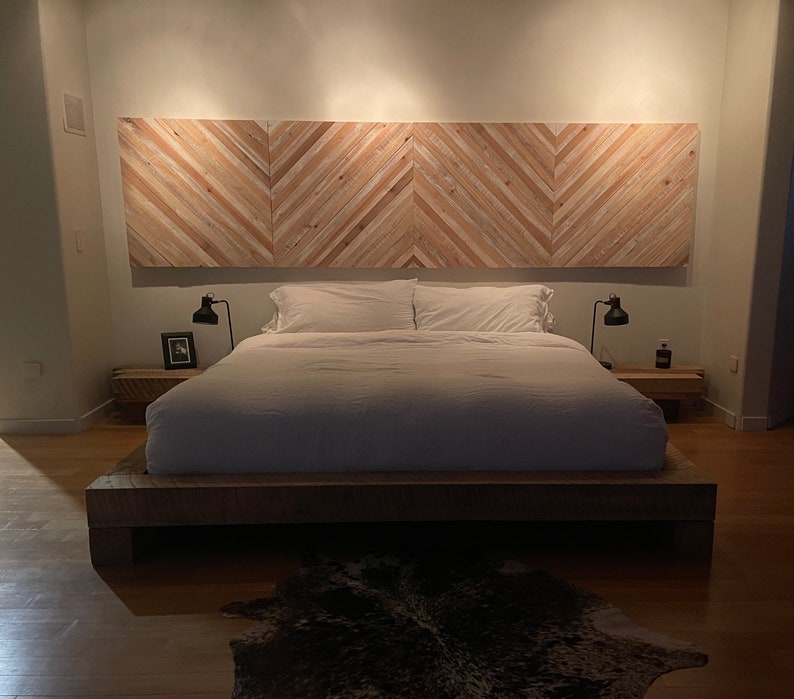 May include: A white bed with a wooden headboard made of light wood planks arranged in a chevron pattern. The bed has a white duvet and two white pillows. There are two bedside tables with lamps on either side of the bed.