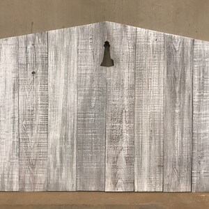 Puede incluir: Se&ntilde;al de madera blanqueada con un recorte de una campana en el centro. La se&ntilde;al est&aacute; hecha de madera recuperada y tiene un aspecto r&uacute;stico.