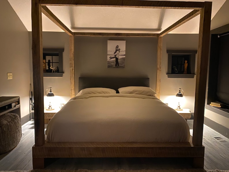 May include: A wooden canopy bed frame with a white duvet and pillows. The bed is in a bedroom with gray walls and a black and white photo on the wall.