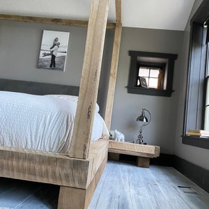 May include: A wooden bed frame with a white bedspread and a gray headboard. The bed frame has a rustic look and is made of thick, unfinished wood. The bed is in a bedroom with gray walls and a small window. There is a nightstand next to the bed with a lamp on it.