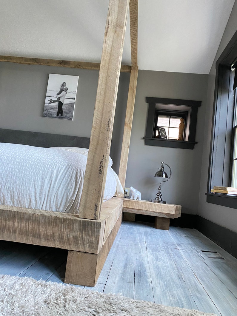 May include: A wooden bed frame with a white bedspread and a gray headboard. The bed frame has a rustic look and is made of thick, unfinished wood. The bed is in a bedroom with gray walls and a small window. There is a nightstand next to the bed with a lamp on it.