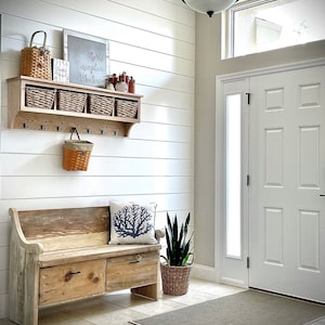 May include: A rustic wooden bench with two drawers and a cushion, a plant in a woven basket, and a shelf with baskets and hooks on a white shiplap wall. The text on the sign reads "There is no place like home".