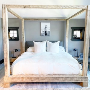 May include: A white bed with a wooden canopy frame and a white duvet. The bed has two white pillows and a black and white photo on the wall behind it.