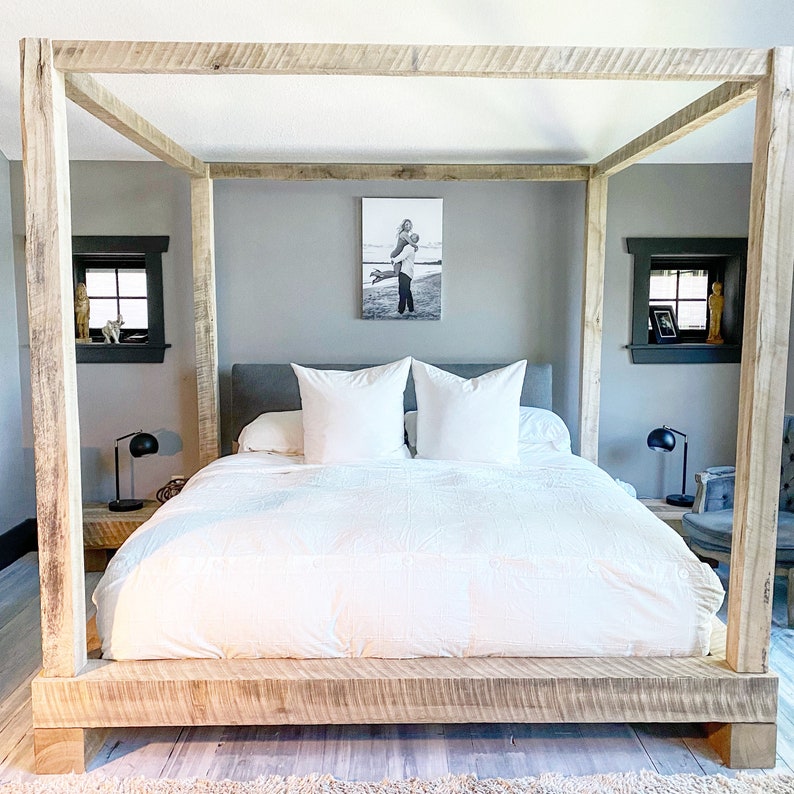 May include: A white bed with a wooden canopy frame and a white duvet. The bed has two white pillows and a black and white photo on the wall behind it.