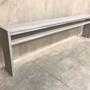 May include: A long, whitewashed wooden console table with two open shelves. The table is standing on a concrete floor.