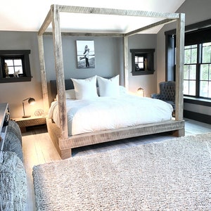 May include: A bedroom with a large wooden canopy bed with white bedding and a gray rug. There is a small table with a lamp next to the bed and a black armchair in the corner.