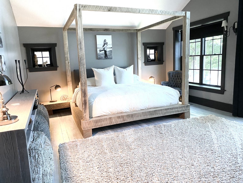 May include: A bedroom with a large wooden canopy bed with white bedding and a gray rug. There is a small table with a lamp next to the bed and a black armchair in the corner.