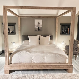 May include: A rustic wooden canopy bed frame with a white duvet and two white pillows. The bed is in a bedroom with a gray wall and a framed photo above the headboard.