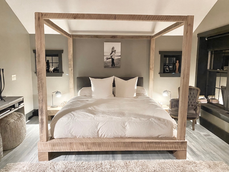 May include: A rustic wooden canopy bed frame with a white duvet and two white pillows. The bed is in a bedroom with a gray wall and a framed photo above the headboard.