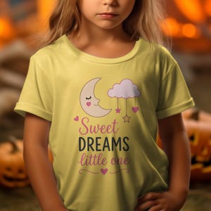 Puede incluir: Una camiseta de color amarillo pálido con un gráfico de una luna creciente dormida, una nube rosa y estrellas colgantes. El texto "Sweet DREAMS little one" está impreso en una mezcla de letras rosas y gris oscuro, con pequeños corazones rosas.