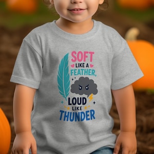 Puede incluir: Una camiseta gris jaspeado de manga corta con un gráfico que incluye una pluma verde azulado, una nube de tormenta gris oscuro con cara de enfado y un rayo amarillo. El texto dice: SOFT LIKE A FEATHER, LOUD LIKE THUNDER, con corazones y estrellas rosas.